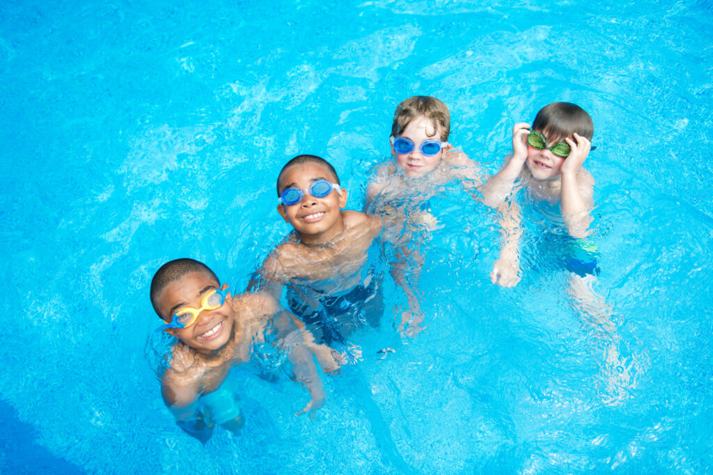 Four young kids wearing swim goggles smile and play together in a bright blue swimming pool, looking up toward the camera.