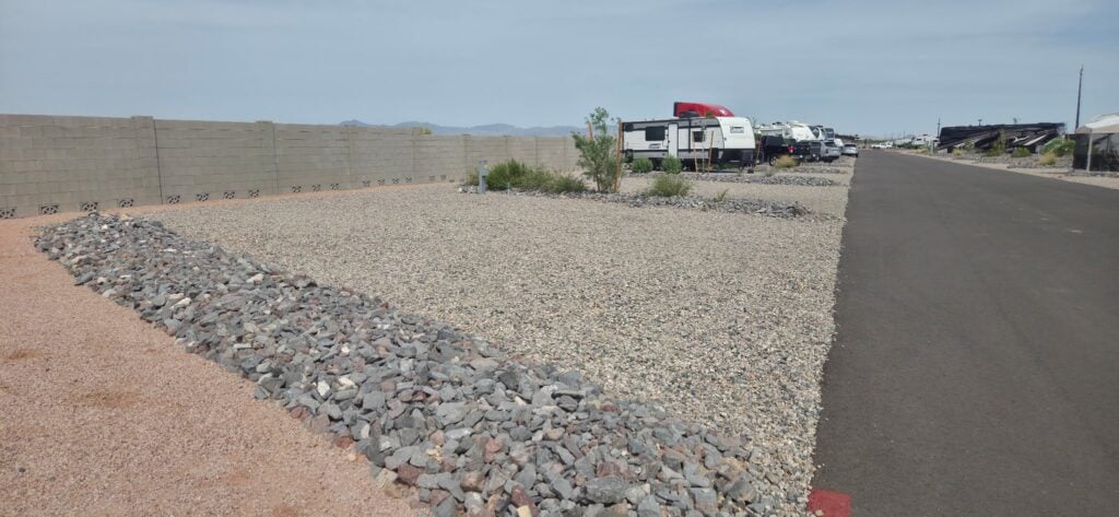 A gravel lot with a small bush and rock border sits beside an asphalt road in an RV park, with several RVs parked along the road and a concrete block wall on the left. Distant mountains are visible under a clear sky.