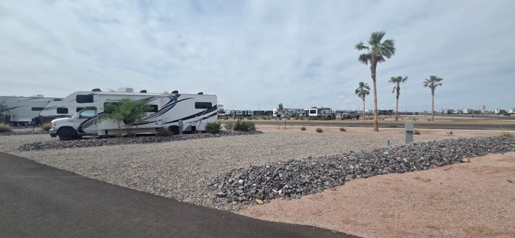 A white recreational vehicle (RV) is parked on a gravel lot in a mostly empty RV park, with palm trees and other RVs in the background under a partly cloudy sky.