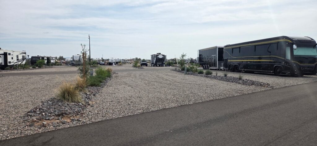 A wide RV park with several large RVs parked on gravel lots, separated by small landscaped islands with rocks and sparse vegetation, under a cloudy sky.