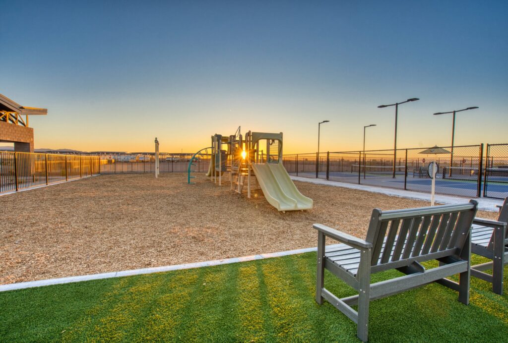 A playground at sunset with slides, climbing structures, and a mulch surface. A wooden bench sits on artificial grass in the foreground, and the area is enclosed by a black fence.
