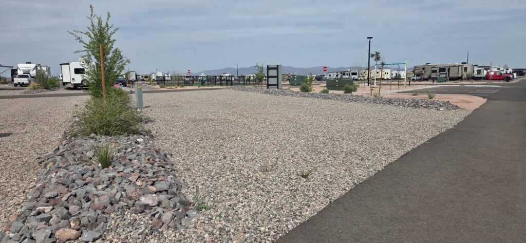RV park with gravel lots, young trees, and small plants. Several RVs are parked in the background. A paved road runs alongside the lots under a partly cloudy sky with mountains visible in the distance.