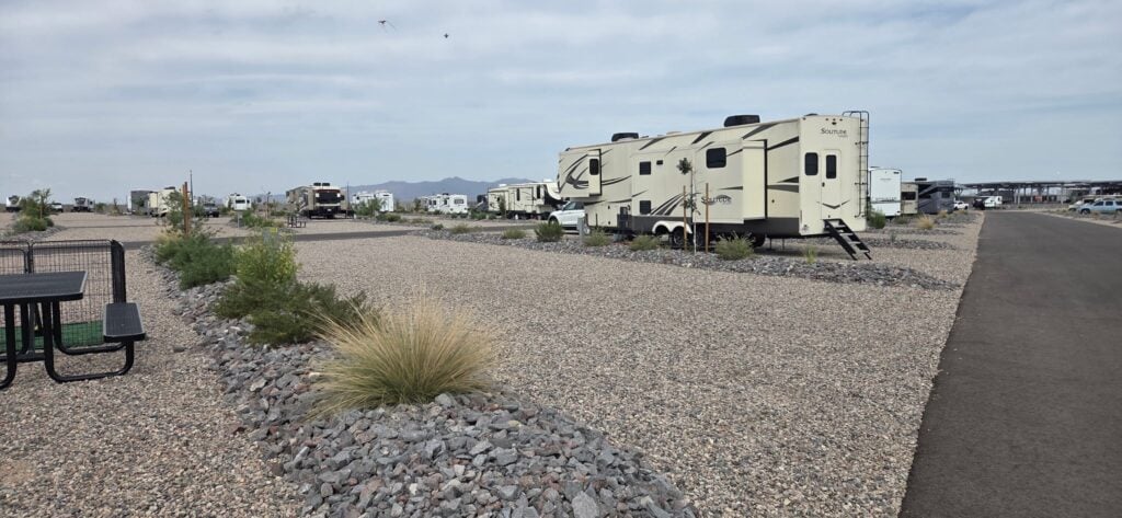 A gravel RV park with several parked RVs, small desert plants along the gravel lots, a picnic table on the left, and an empty lot in the foreground. Mountains are visible in the distant background under a cloudy sky.