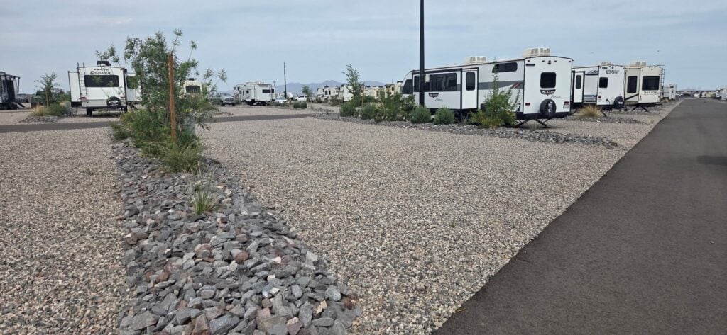 A row of white RVs parked on gravel lots next to a paved road, with small trees and rocks in landscaped strips between the lots under a cloudy sky.