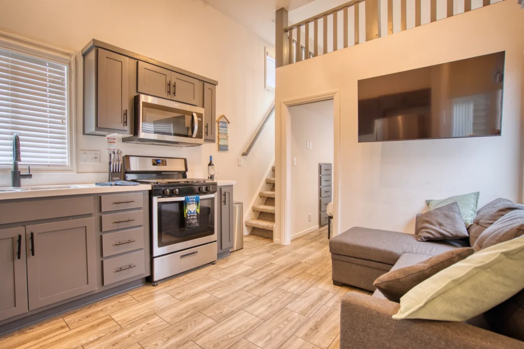 Modern kitchen and living area with gray cabinets, stainless steel appliances, sectional sofa, and a large wall-mounted TV; stairs lead to an upper loft. Light wood flooring and neutral tones create a cozy atmosphere.