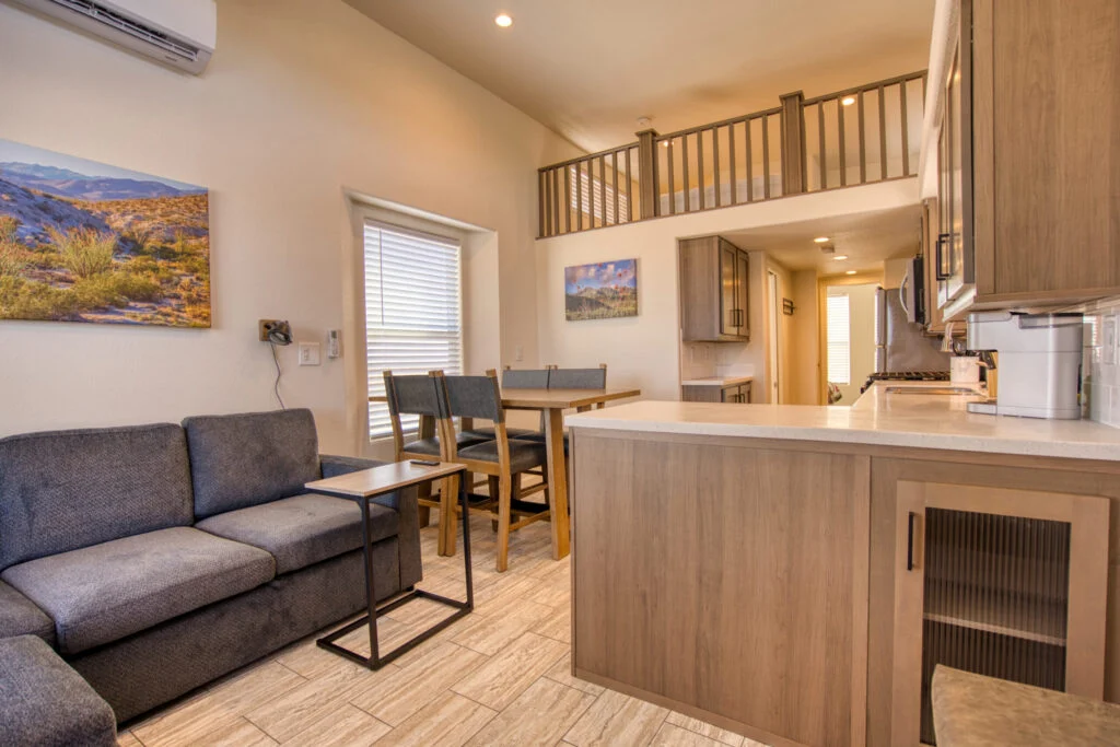 Modern kitchen and living area with a gray couch, dining table, light wood cabinets, wall art, and a loft above. Natural light enters through windows, creating a cozy, inviting atmosphere.