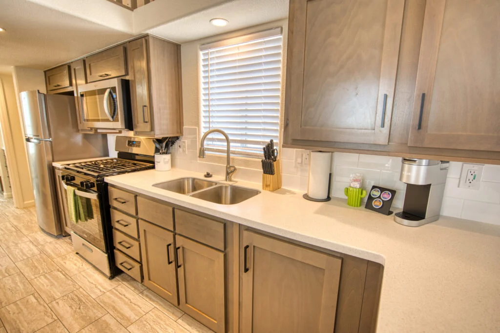 Modern kitchen with light wood cabinets, stainless steel appliances, a double sink under a window with blinds, and various utensils and small appliances on the countertop.