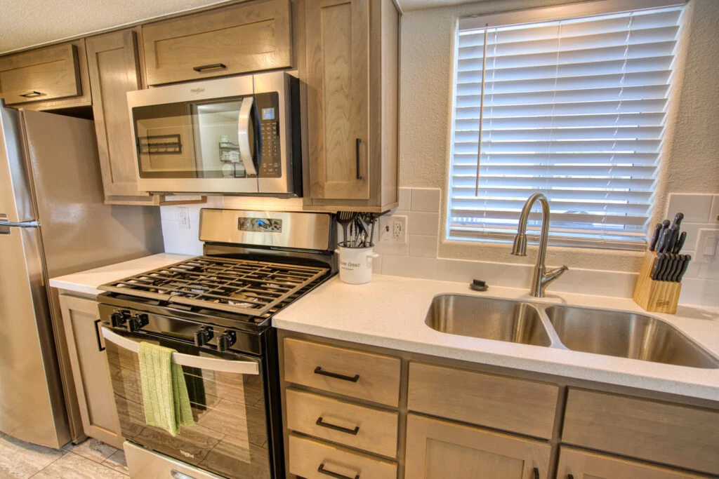 Modern kitchen with stainless steel appliances, gas stove with oven, microwave above, double sink with faucet, wooden cabinets, a knife block, and a window with white blinds. A green towel hangs on the oven handle.